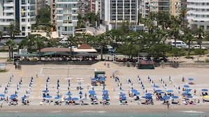 Ein Strand mit Reihen von blauen Sonnenschirmen und Liegestühlen, ein Gebäude mit grüner Dachbegrünung und ein hohes, weißes Gebäude im Hintergrund.