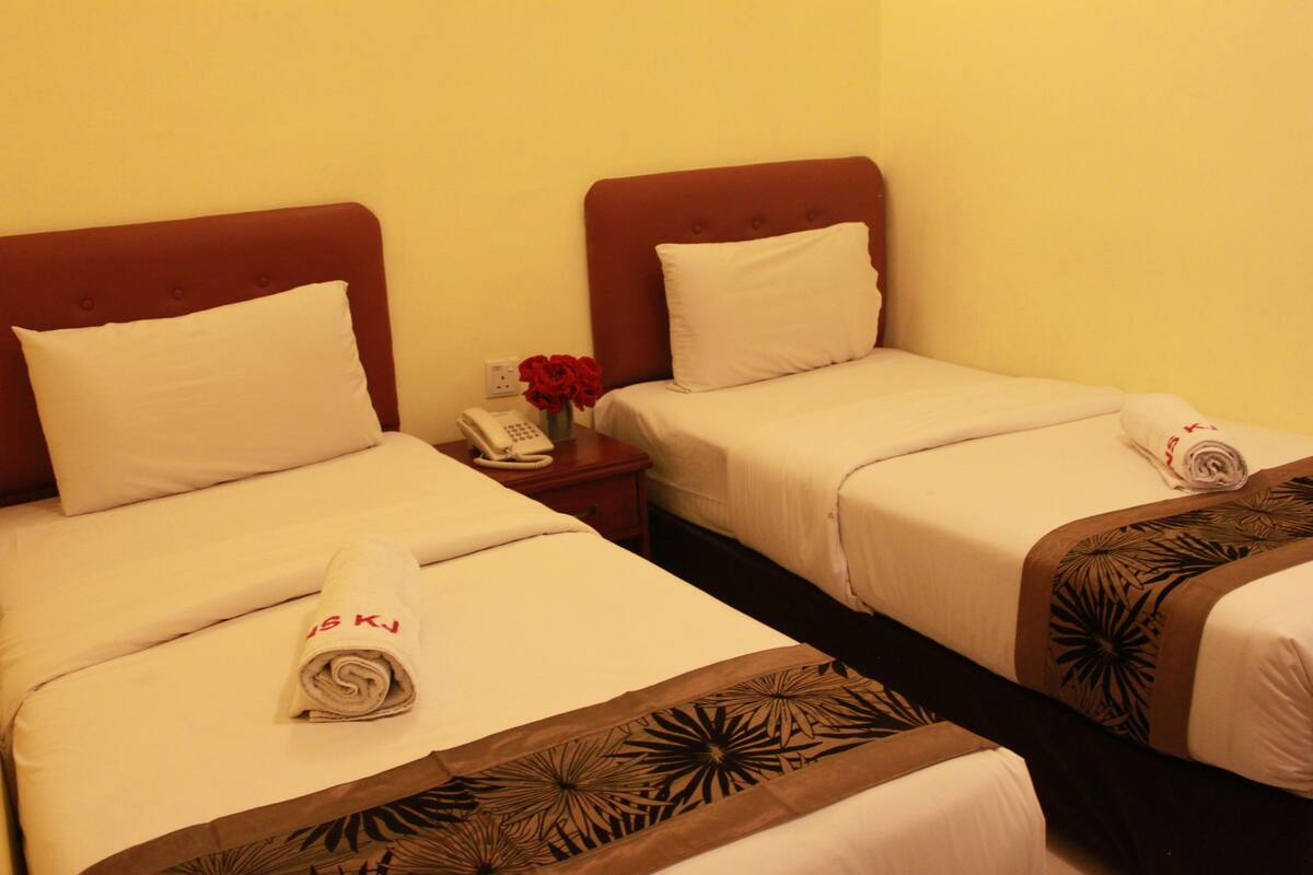 Habitación de hotel con dos camas, cada una con ropa blanca y cabeceros marrones. Hay un teléfono en la mesita de noche y un jarrón con flores rojas.