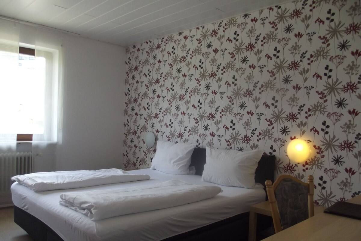 Ein Schlafzimmer mit Bett, Nachttisch, Lampe, Fenster und Heizkörper.