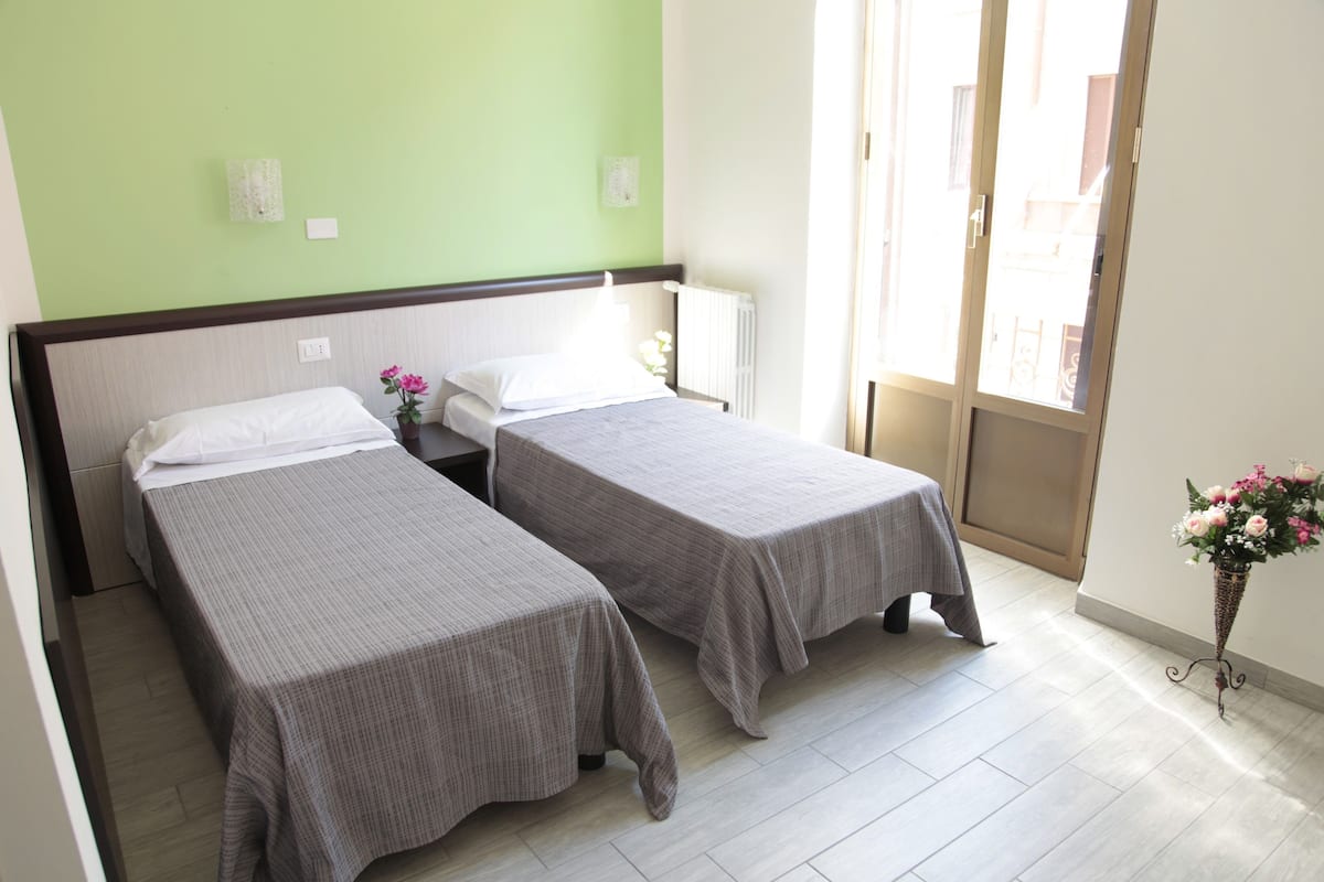 A hotel room with two single beds, a small table, a nightstand, a window with a view, and a vase with flowers.