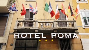Eine Hotelfassade mit mehreren Flaggen, darunter die Vereinigtes Königreich, Italien und weitere, und einem Schild mit der Aufschrift 'HOTEL ROMA' und drei Sternen.