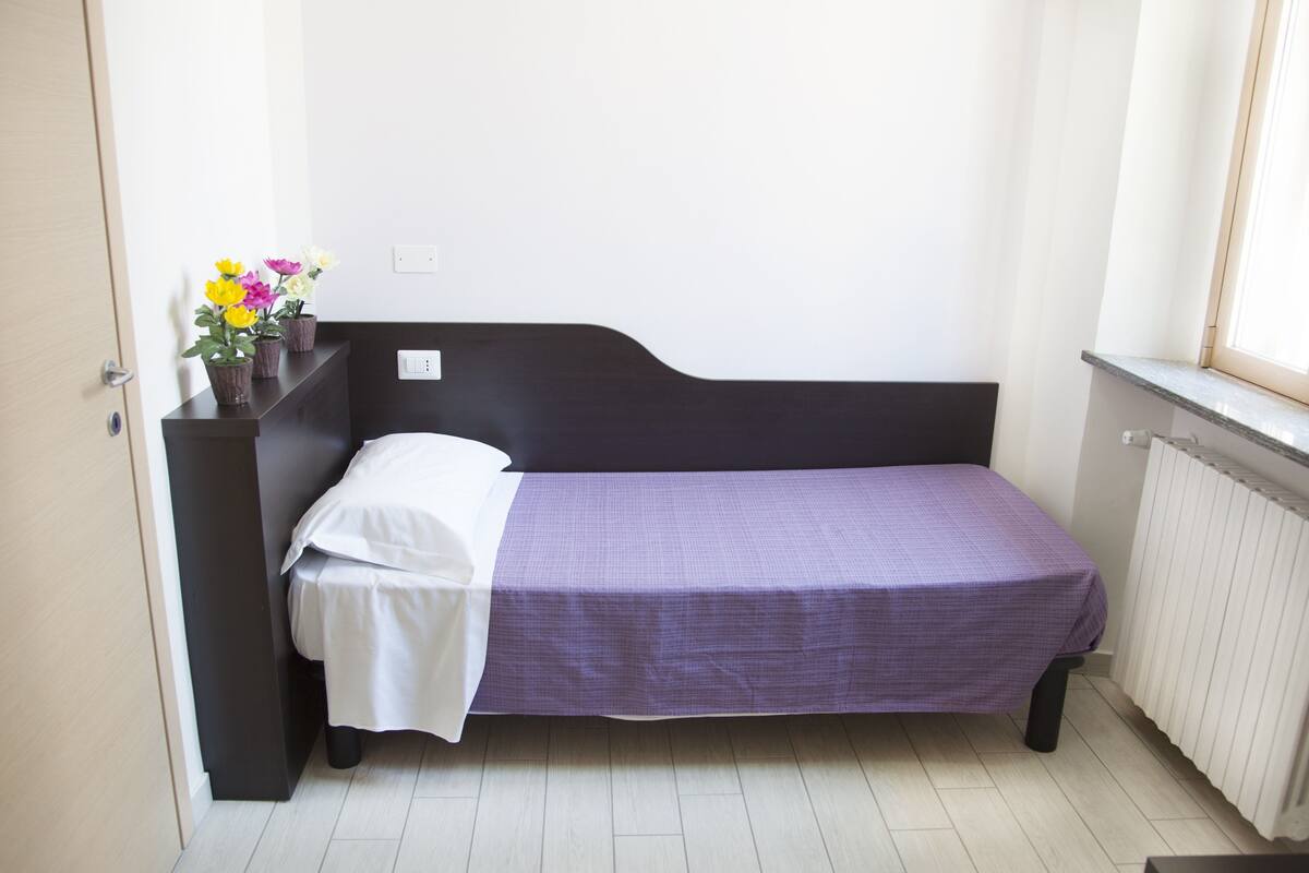 A single bed with a black headboard, a white pillow, and a purple bedspread in a room with white walls and a window.