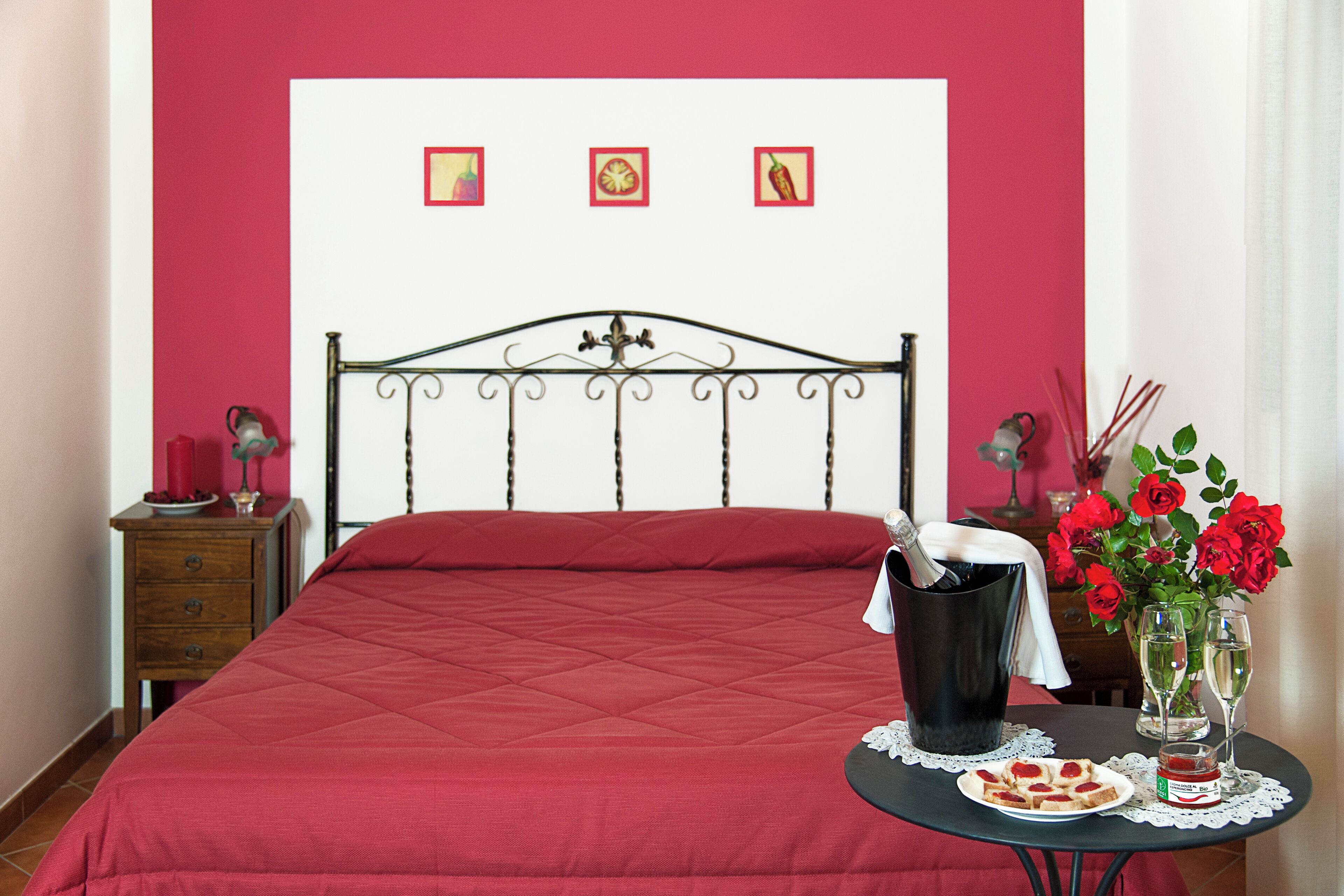 Un letto con una coperta rossa, un comodino con una bottiglia, un vaso con fiori e un piatto con cibo.