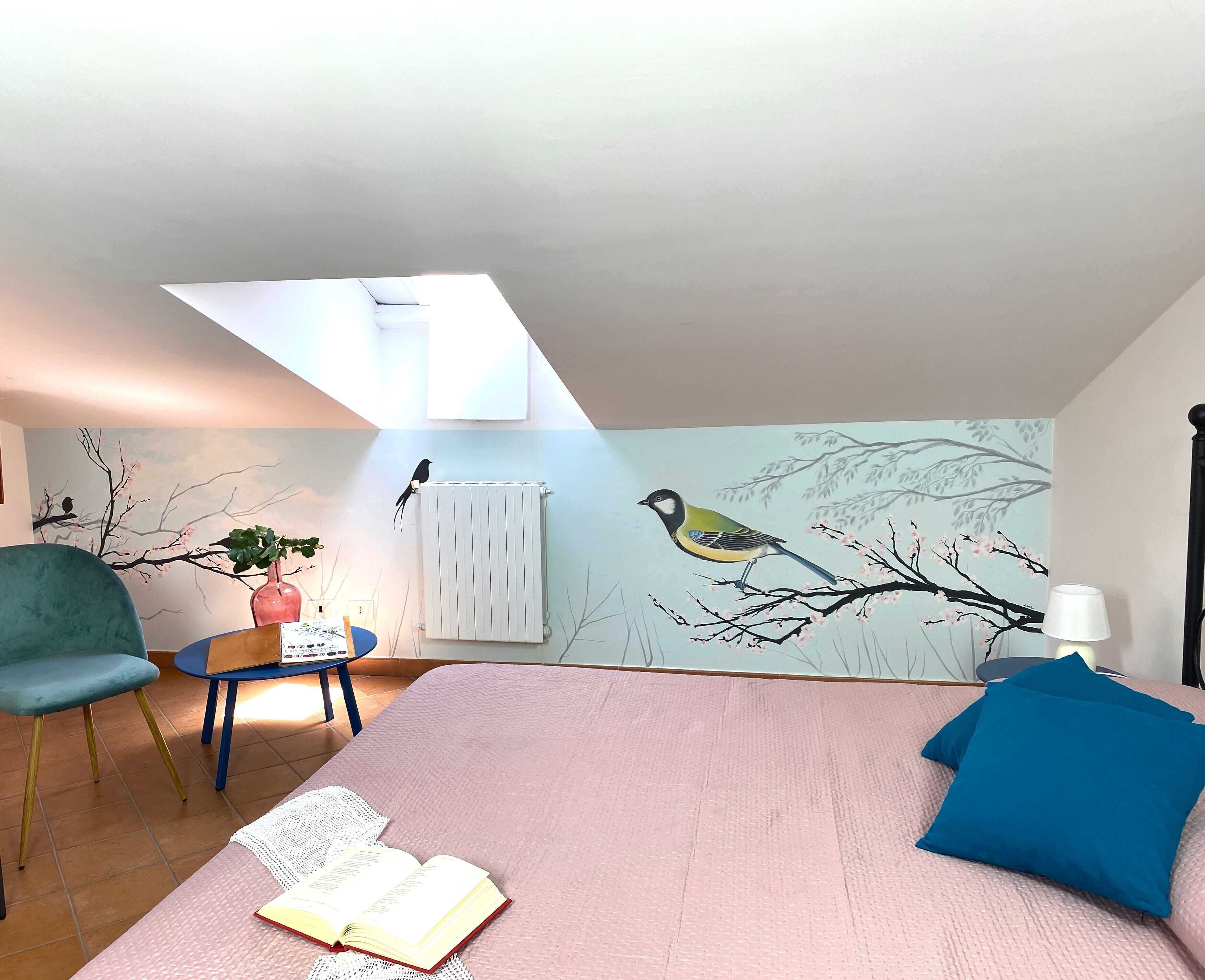 Une chambre avec un plafond en pente, un lit avec une literie rose, une chaise bleue, une petite table avec une plante et une fresque murale représentant des oiseaux perchés sur une branche.