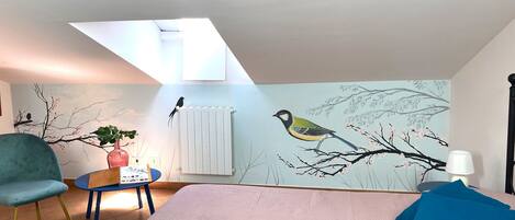Une chambre avec un plafond en pente, un lit avec une literie rose, une chaise bleue, une petite table avec une plante et une fresque murale représentant des oiseaux perchés sur une branche.