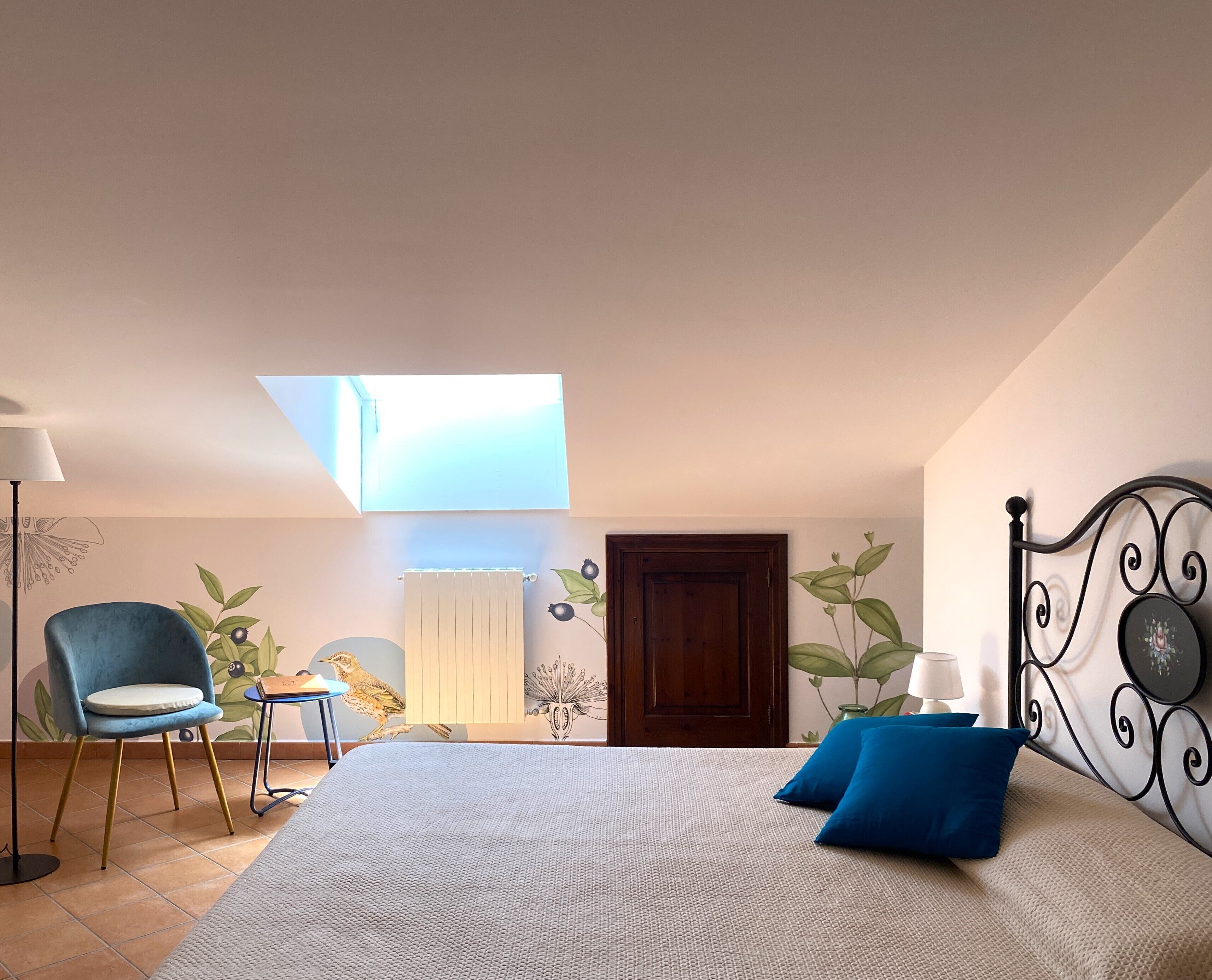 A bedroom with a skylight, a bed with blue pillows, a chair, a small table, and a wall with plant illustrations.
