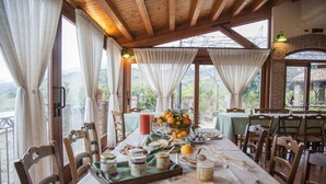 A dining table set with food and drinks, surrounded by wooden chairs, in a room with large windows and a wooden ceiling. - Agriturismo Bergi (Castelbuono)