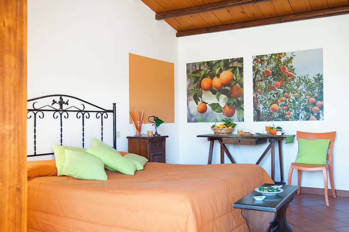 Quarto com cama, mesa com frutas e uma cadeira, e duas pinturas na parede.