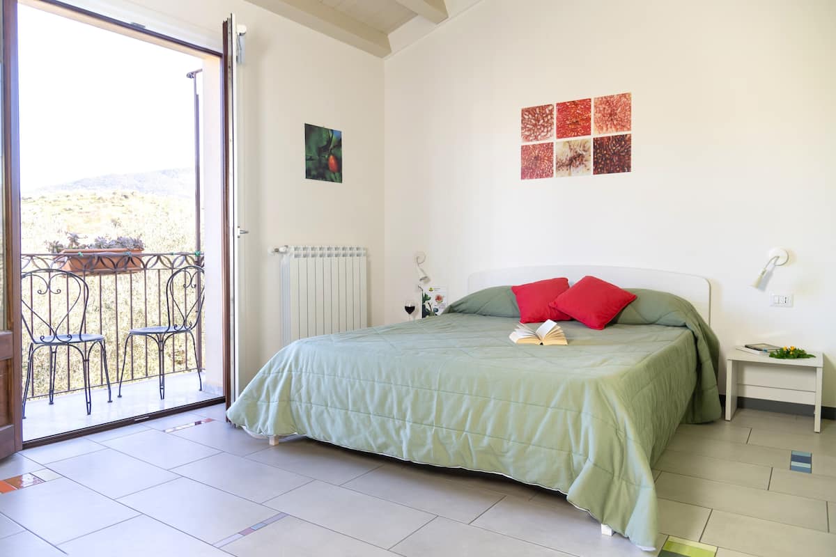 Quarto com cama, mesa de cabeceira, janela com vista para o exterior e porta que dá para uma varanda.