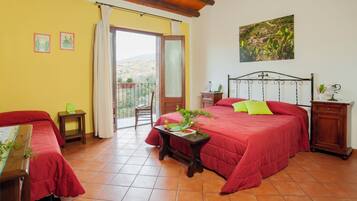 A bedroom with a red bed, a wooden headboard, a small table with a plant, and a balcony with a view of the countryside.