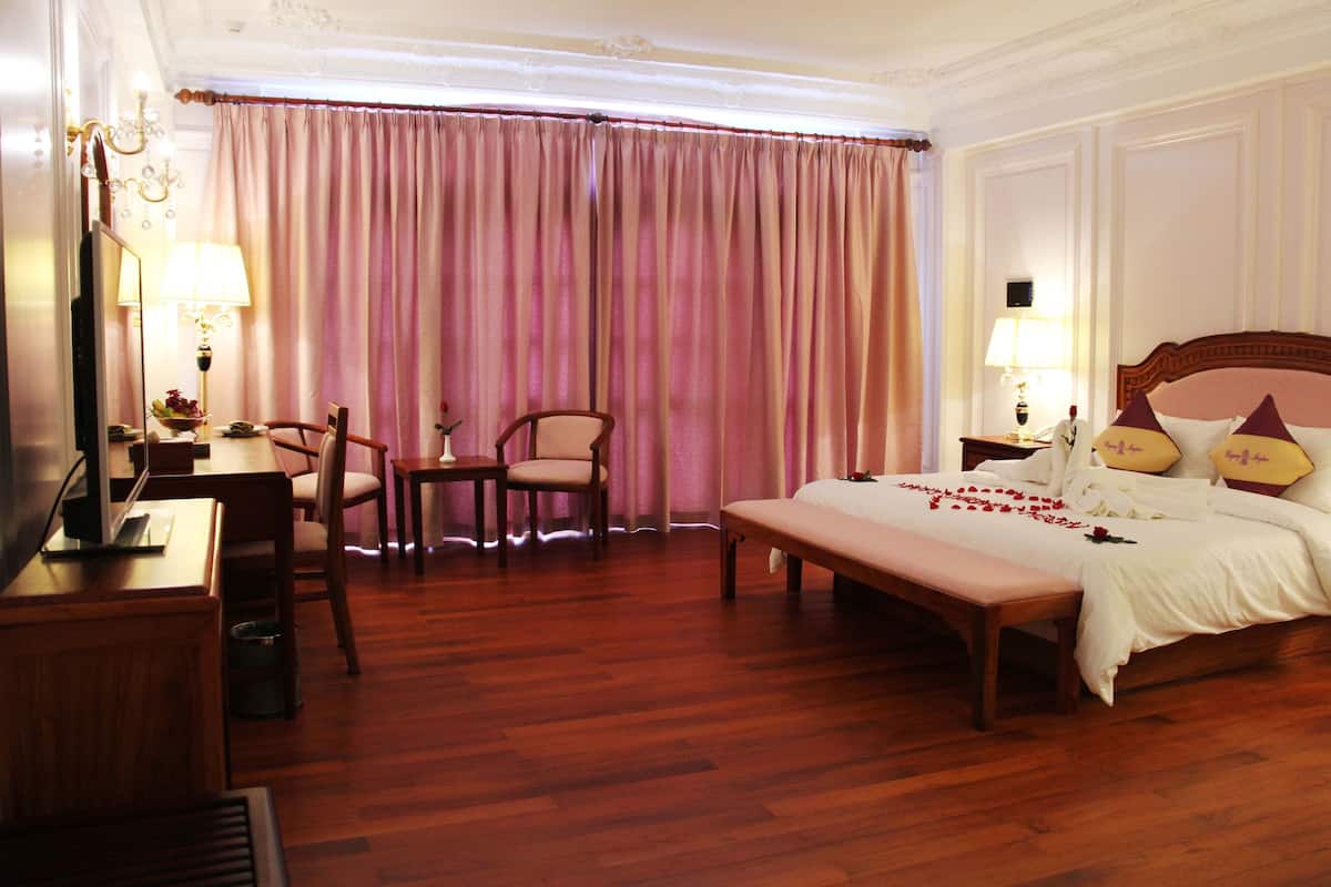 A hotel room with a wooden floor, a bed with a white bedspread and decorative pillows, a desk with a lamp, a chair, and a large window with pink curtains.