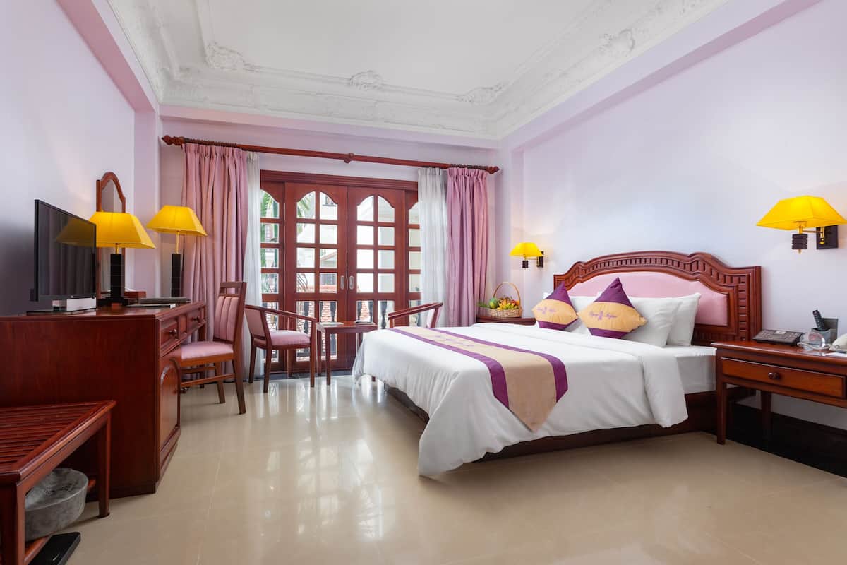 A hotel room with a bed, a desk with a TV, a chair, a lamp, and a window with curtains.