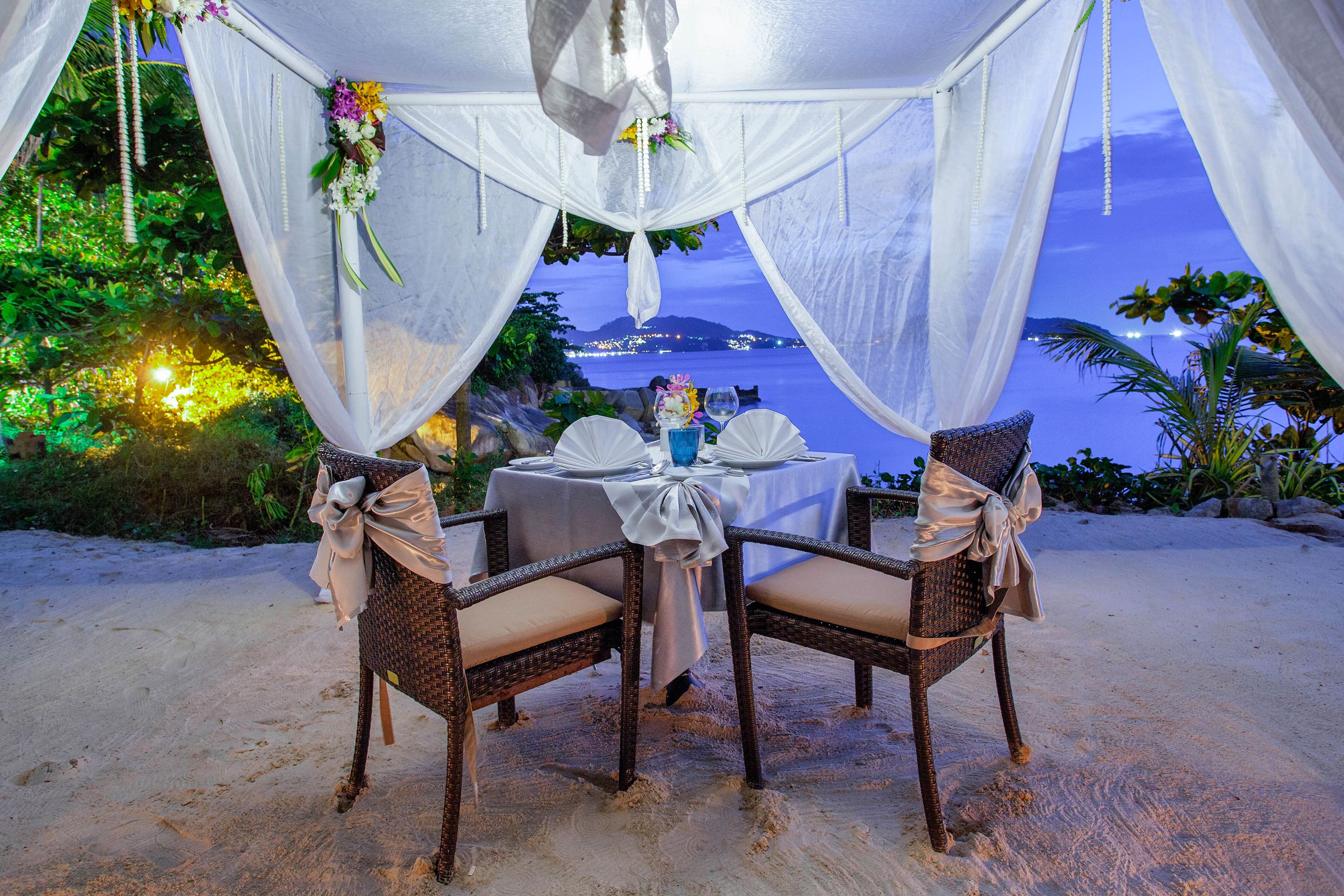 A beachside dining setup with a table and chairs, draped with white fabric and adorned with flowers.