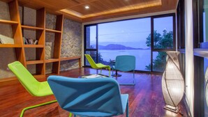 Una habitación moderna con suelo de madera, pared de piedra y un amplio ventanal con vistas a un cuerpo de agua. Hay dos sillas y una estantería.