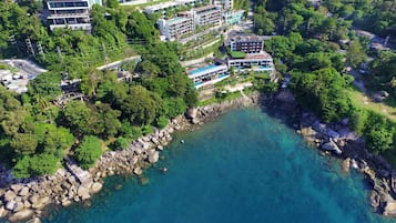 Vista aérea de un complejo turístico costero con varias edificaciones, una costa rocosa y agua azul cristalina.