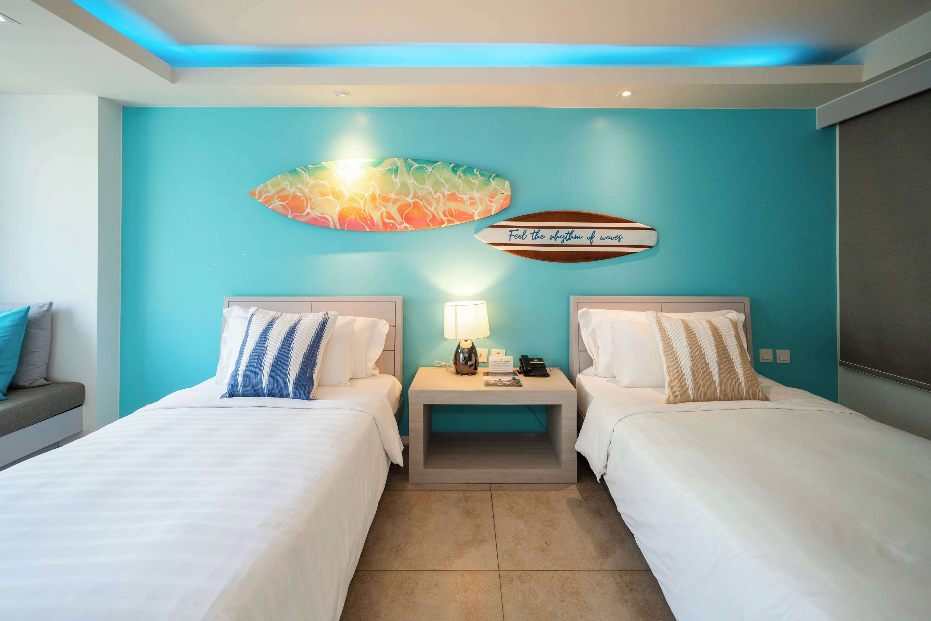 Une chambre d’hôtel avec deux lits, une table de chevet avec une lampe, une œuvre d’art murale et un panneau indiquant : « Ressentez le rythme des vagues. »