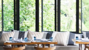 Un comedor con una mesa y sillas de madera, un sofá con cojines a rayas y vistas a la vegetación a través de amplias ventanas.