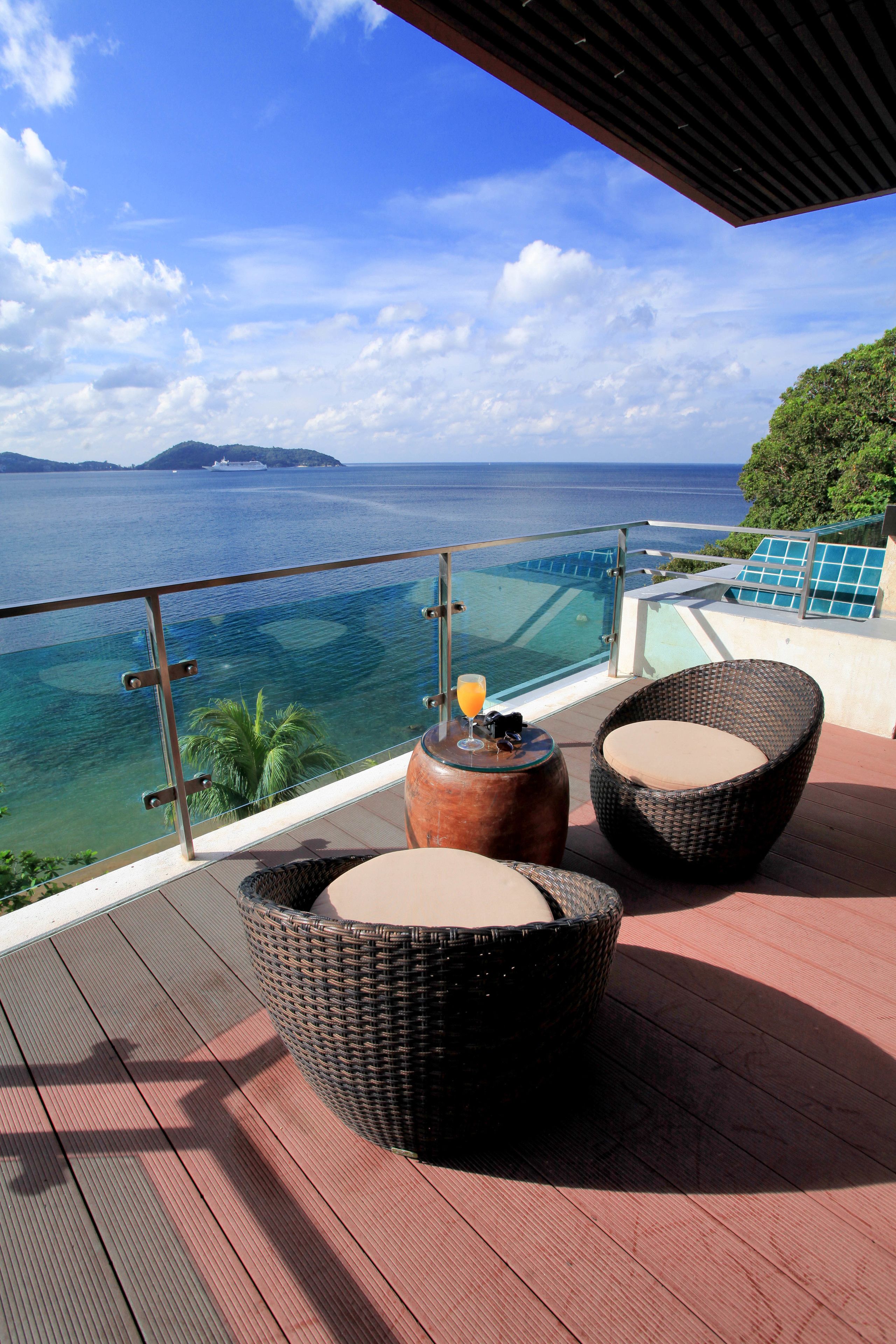 A balcony with wicker chairs and a table, overlooking a clear blue sea and distant islands.