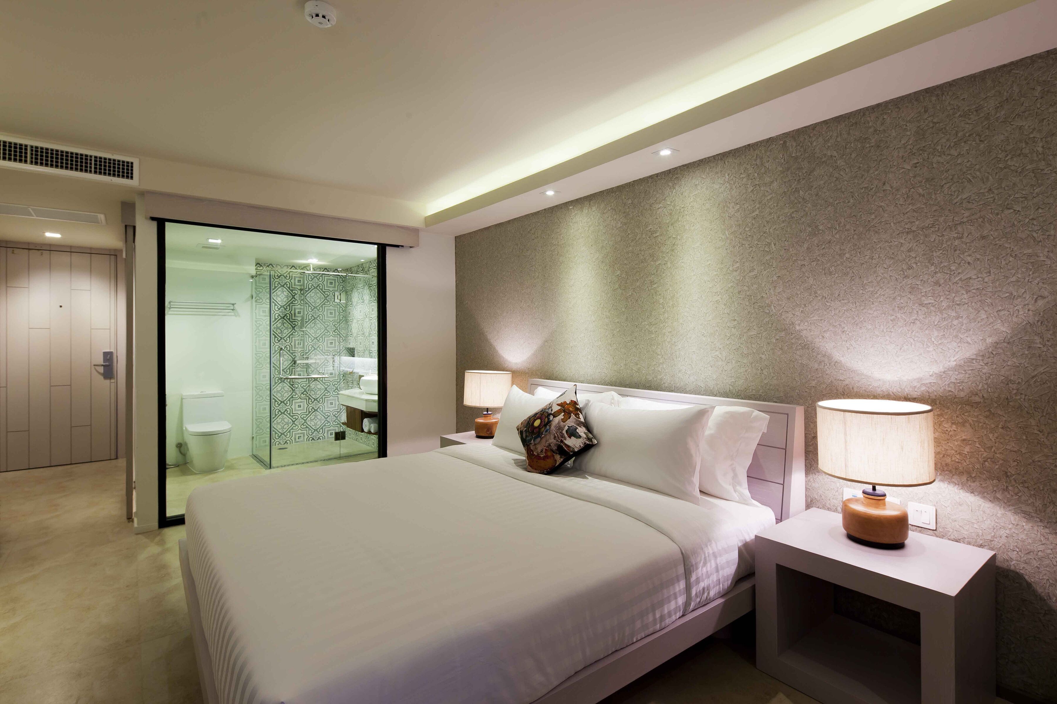 A modern hotel room with a large bed, a nightstand with a lamp, a glass shower enclosure, and a textured wall.