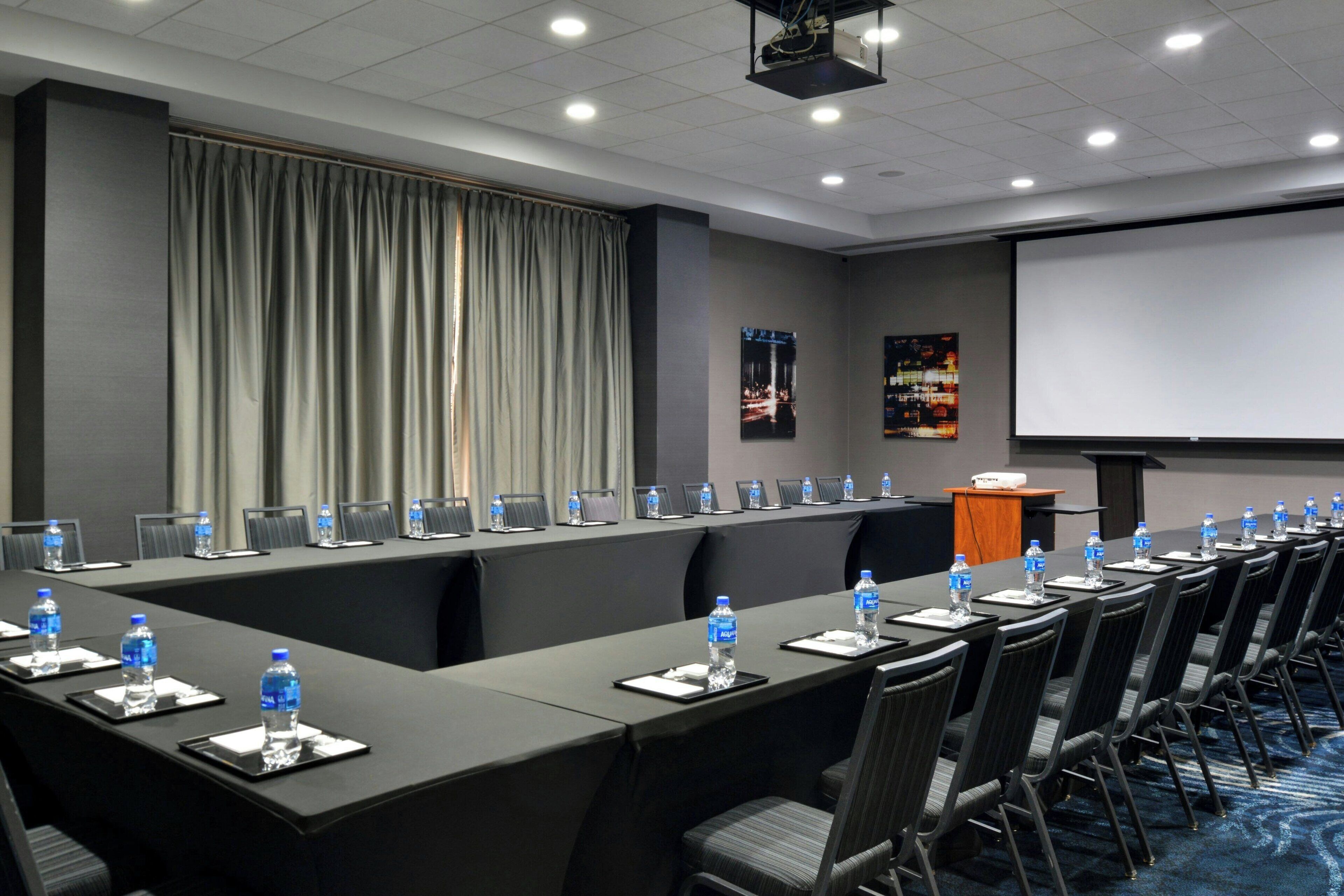 Un salón de conferencias con una larga mesa, sillas, botellas de agua y una pantalla de proyector.