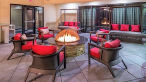 A lounge area with wicker chairs and red cushions, a central fire pit, and a view of a fire through large windows. - Courtyard by Marriott Wilmington Downtown/Historic District (Wilmington)