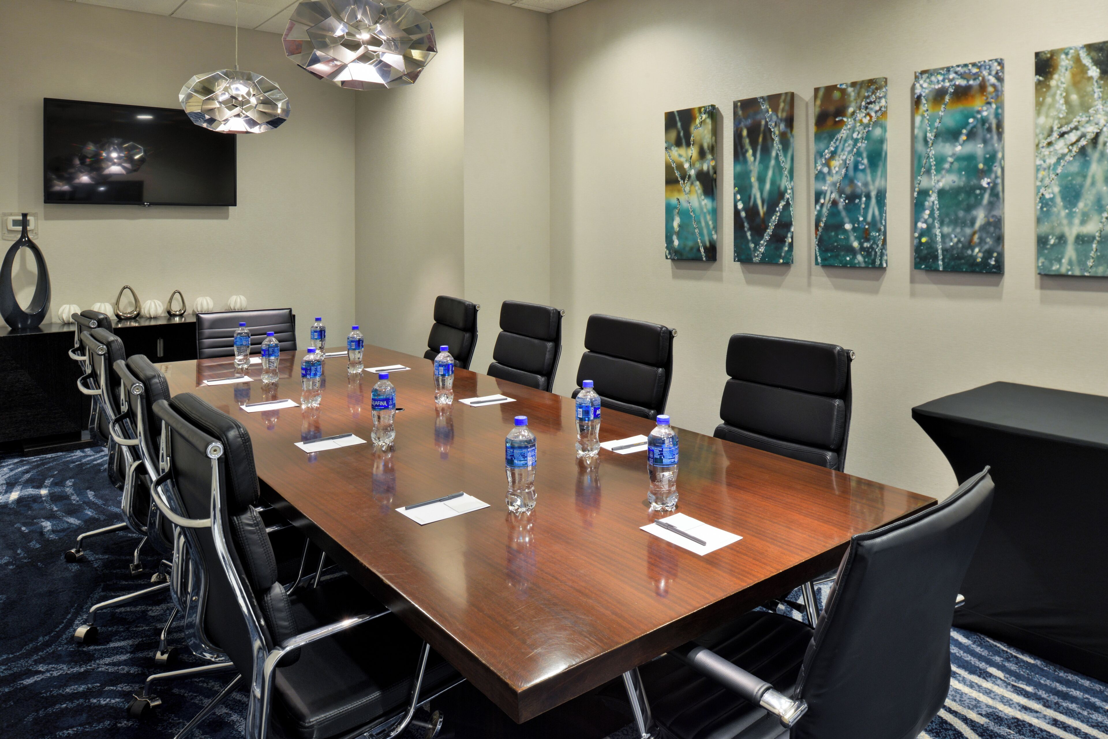 A conference room with a long wooden table, black chairs, water bottles, and a TV on the wall.