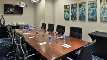 A conference room with a long wooden table, black chairs, water bottles, and a TV on the wall.
