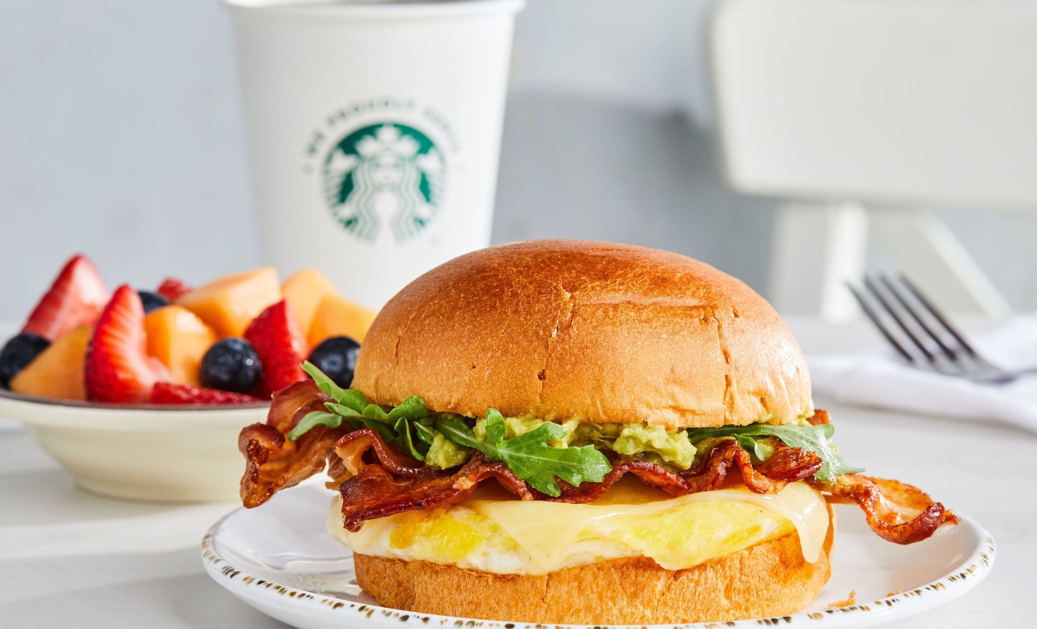 Un sándwich de desayuno con tocino, lechuga y queso en un pan, acompañado de un bol de fruta y una taza de Starbucks.
