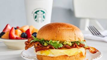 Un sándwich de desayuno con tocino, lechuga y queso en un pan, acompañado de un bol de fruta y una taza de Starbucks.