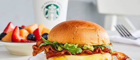 A breakfast sandwich with bacon, lettuce, and cheese on a bun, accompanied by a fruit bowl and a Starbucks cup.