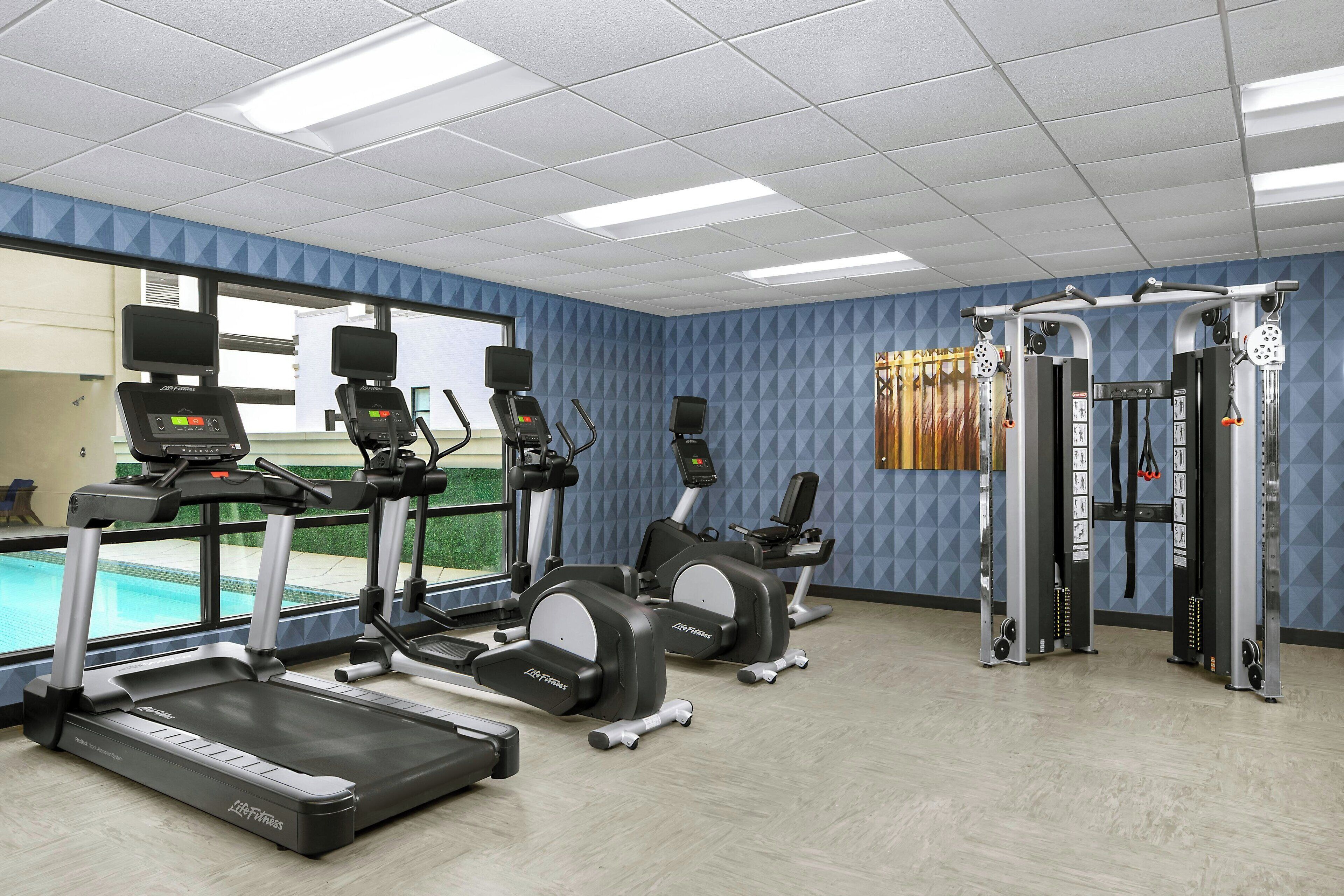 Un gimnasio con cintas de correr, elípticas y máquinas de pesas, amplias ventanas y vista a una piscina.