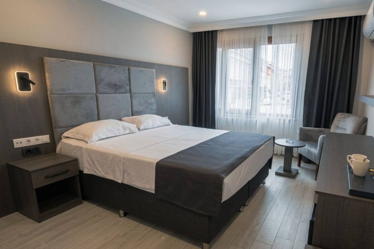 Une chambre d’hôtel moderne, dotée d’un grand lit, de tables de chevet, d’un petit bureau et offrant une vue sur la ville grâce à de grandes fenêtres.