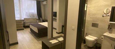 Una habitación de hotel moderna con una cama, un escritorio pequeño, un espejo y un baño con inodoro y lavabo.