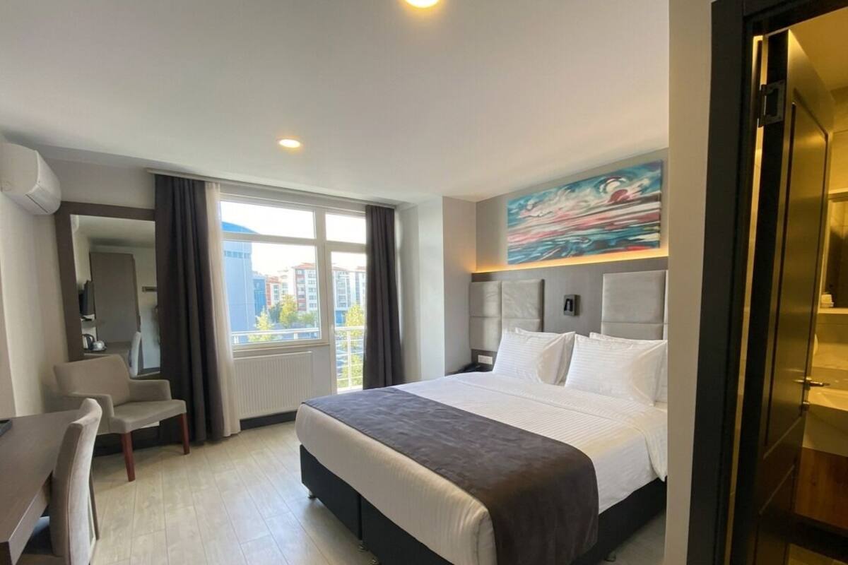 Une chambre d’hôtel avec un lit, un bureau, une chaise, une fenêtre avec des rideaux et un tableau au mur.