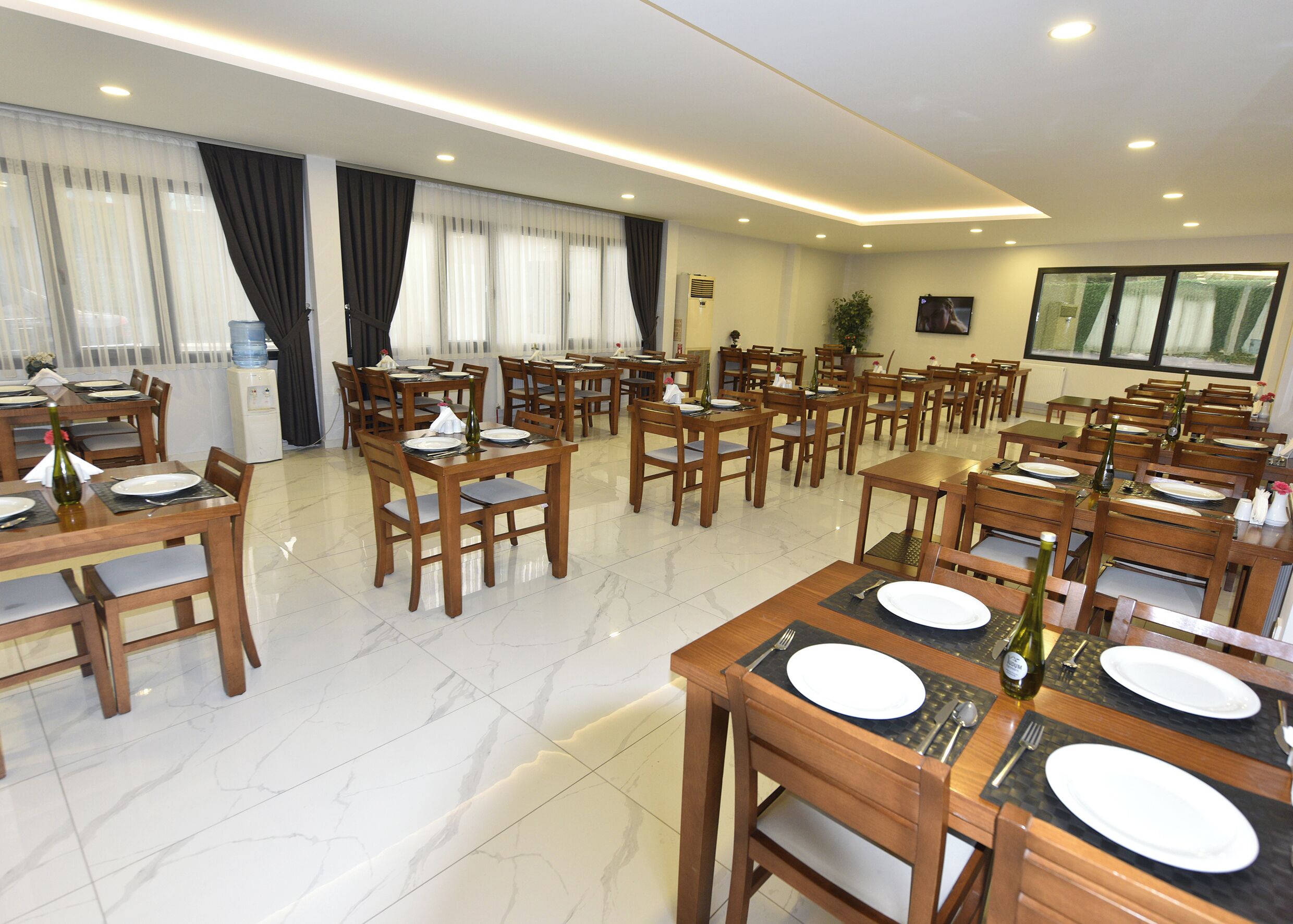 Une salle à manger d’hôtel avec plusieurs tables et chaises, un distributeur d’eau et une grande fenêtre avec des rideaux.