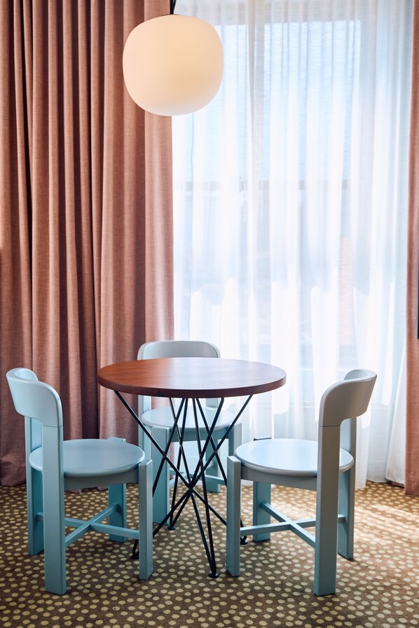 A room with a round table, four chairs, a hanging light fixture, and a window with curtains.