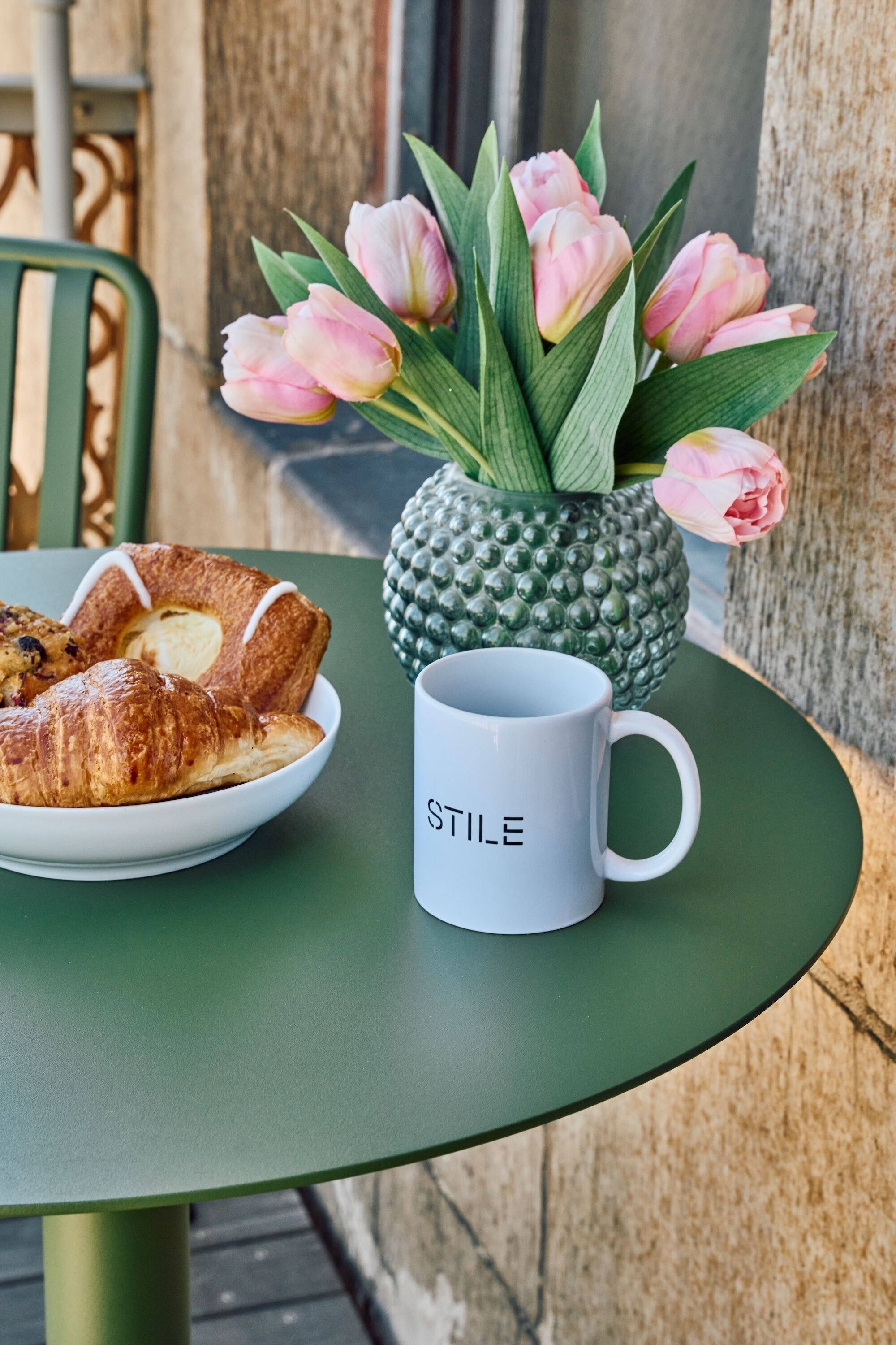 Una mesa con un plato de croissants, un florero con forma de piña y tulipanes rosados, y una taza con la palabra 'STILE'.