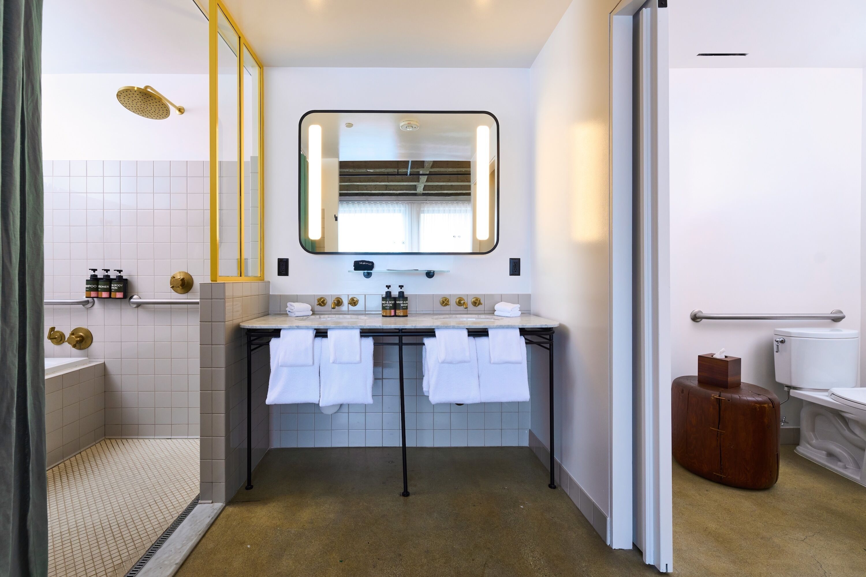 A bathroom with a shower, two sinks, a large mirror, and a suitcase.