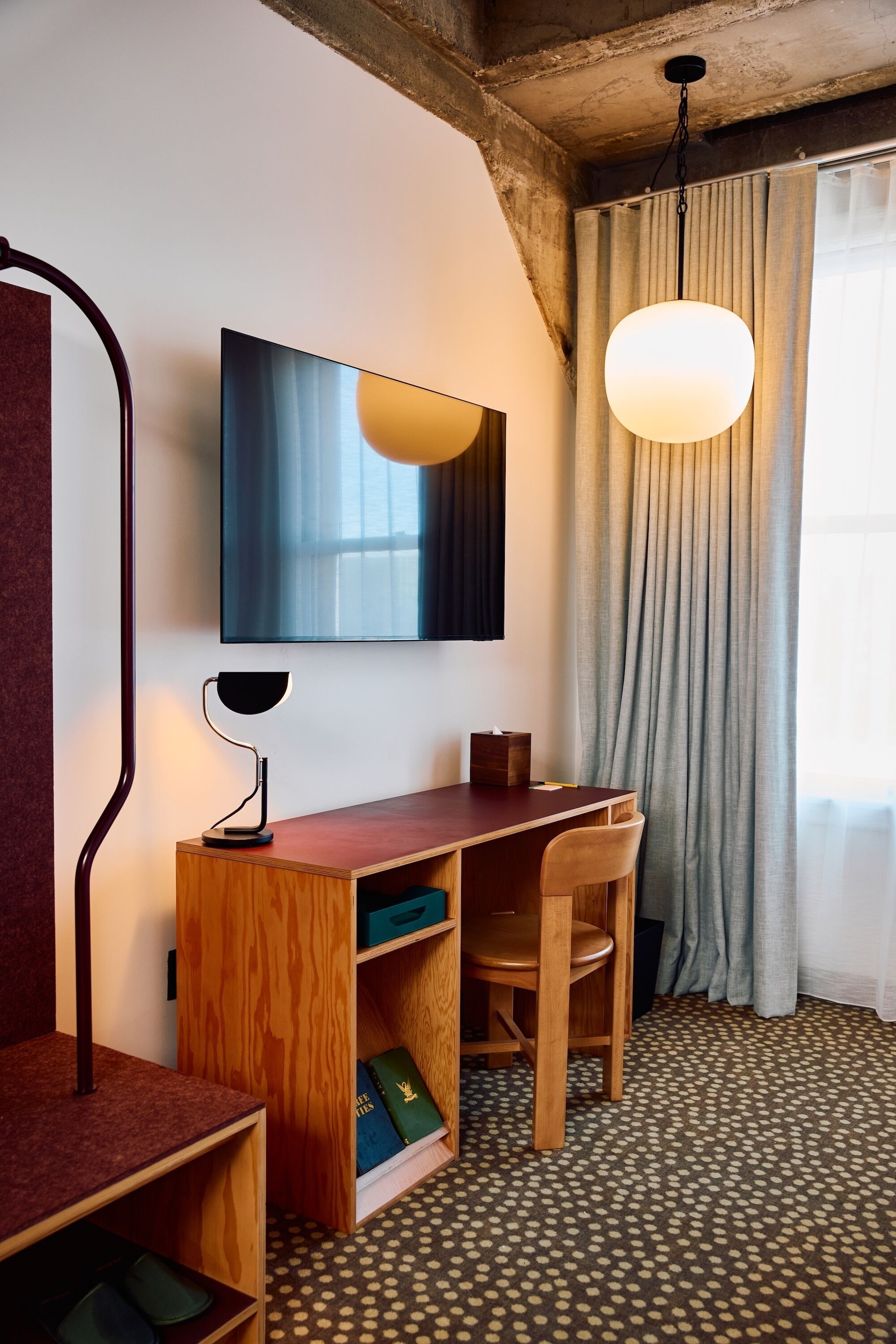 A room with a desk, chair, books, a lamp, a hanging light, and a window with curtains.