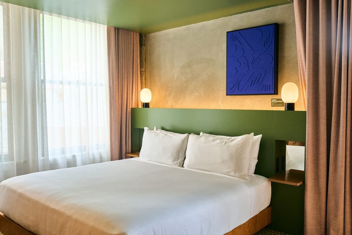 Hotellihuone, jossa on suuri sänky, kaksi yöpöytävalaisinta, seinälle ripustettu taideteos ja ikkuna, jossa on läpikuultavat verhot.
