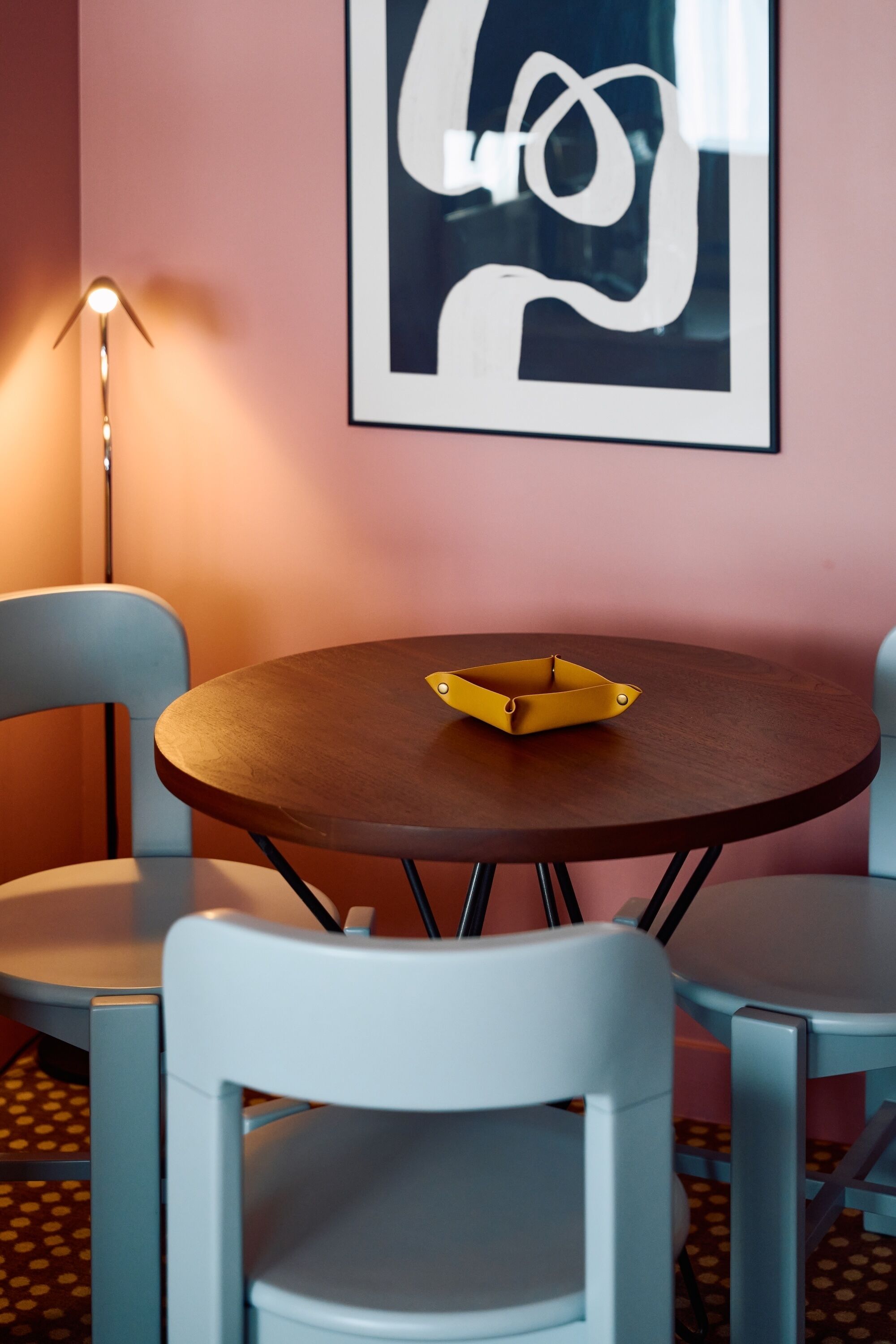 Una mesa redonda de madera con una bandeja amarilla, dos sillas blancas, una lámpara de pie y una obra de arte abstracta enmarcada en la pared.
