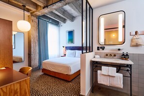 A hotel room with a bed, a desk, a mirror, a bathroom with a sink and toiletries, and a view of the outside.