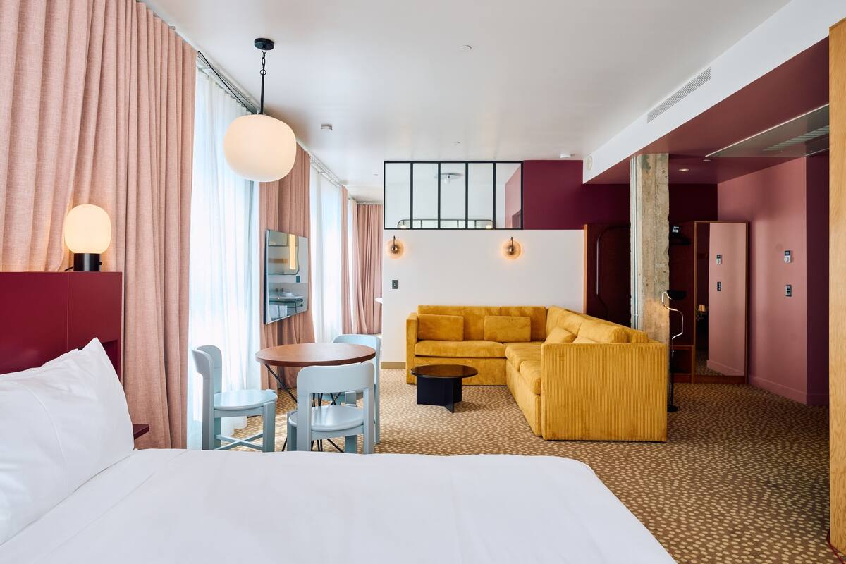 Hotellihuone, jossa on sänky, keltainen sohva, pieni pyöreä pöytä ja ruokailutila tuoleineen.