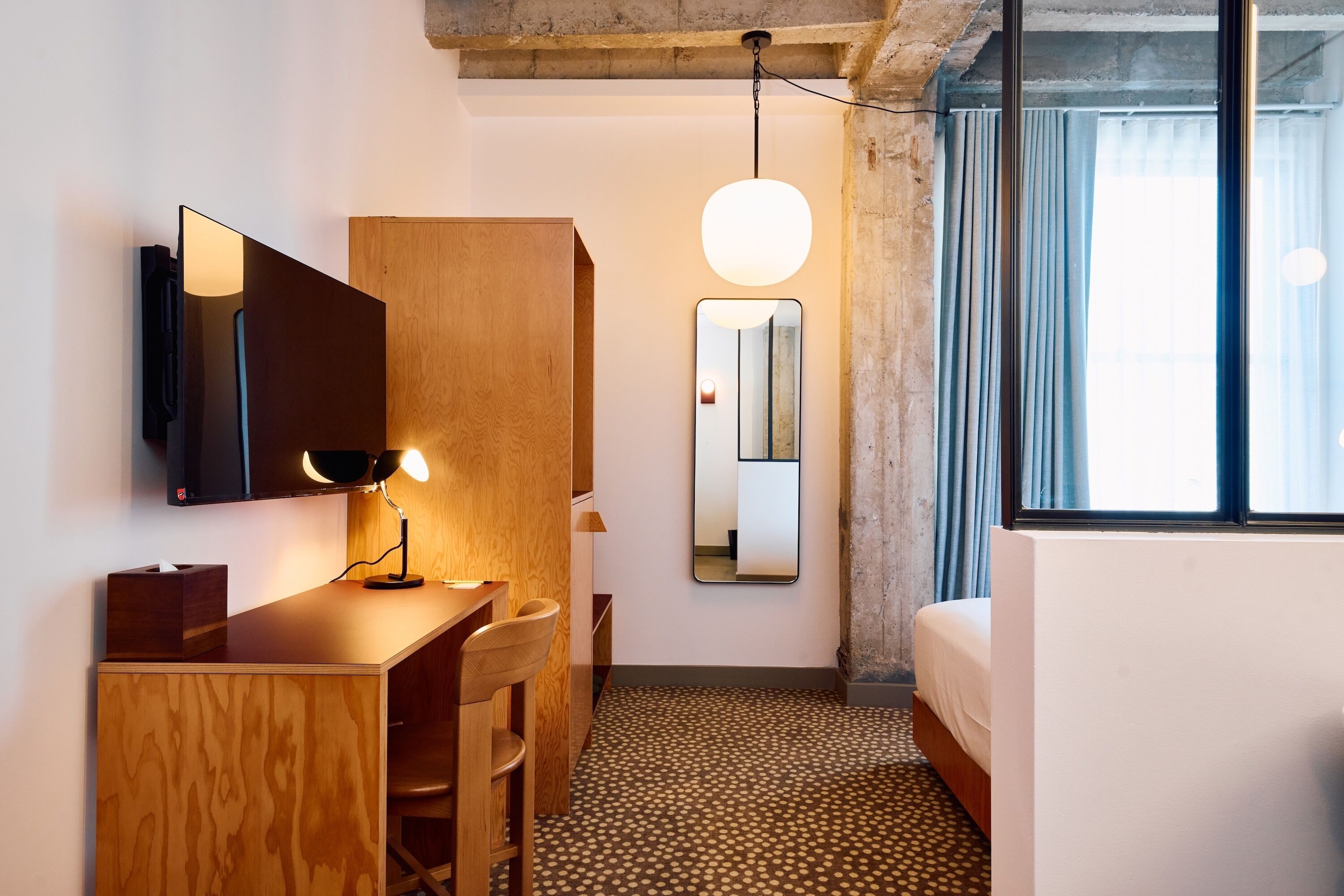A compact hotel room with a wooden desk, a mirror, a bed, and a large window with curtains.