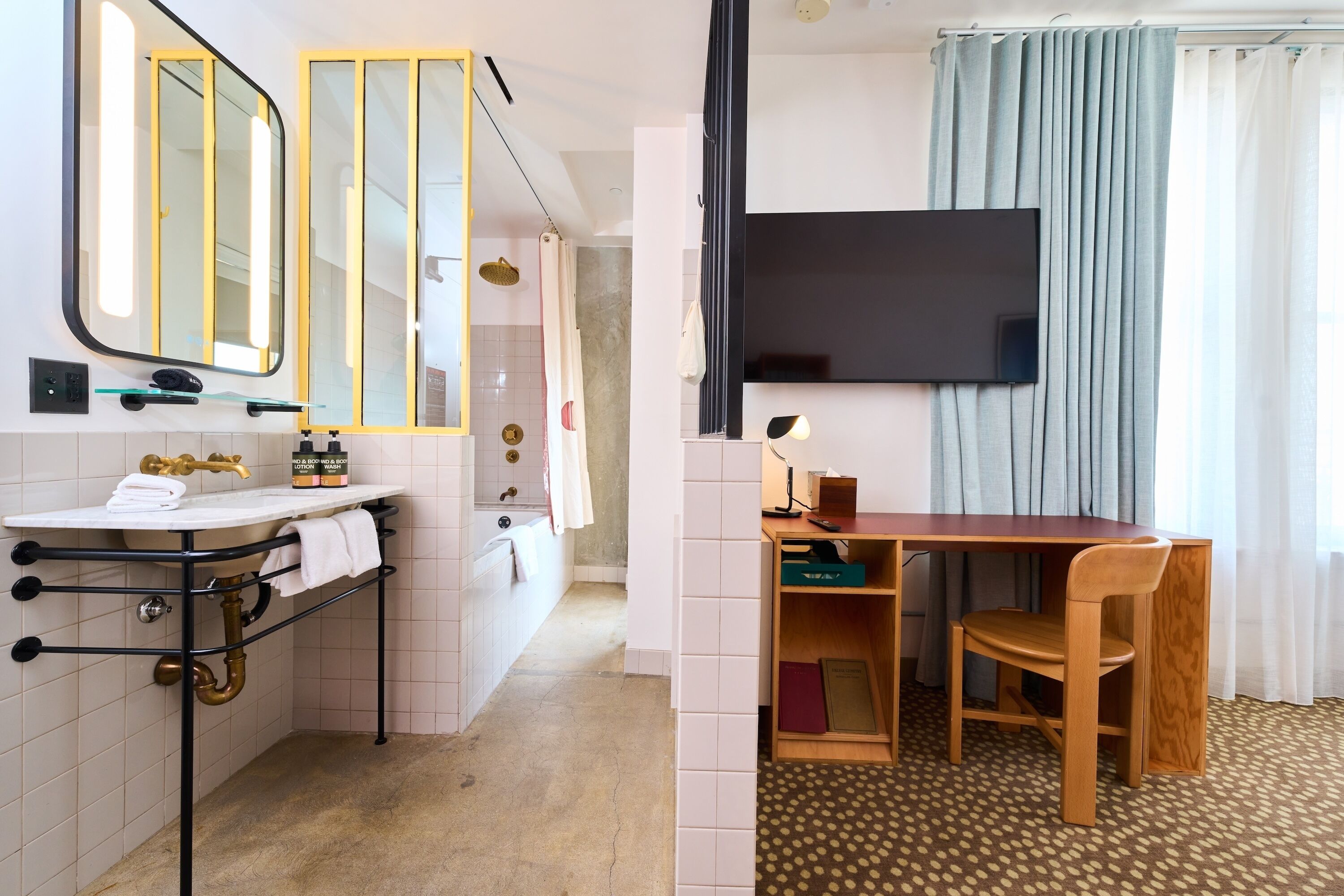 A bathroom with a sink, mirrors, and a shower. There's a TV mounted on the wall and a wooden table with a chair in the adjacent room.
