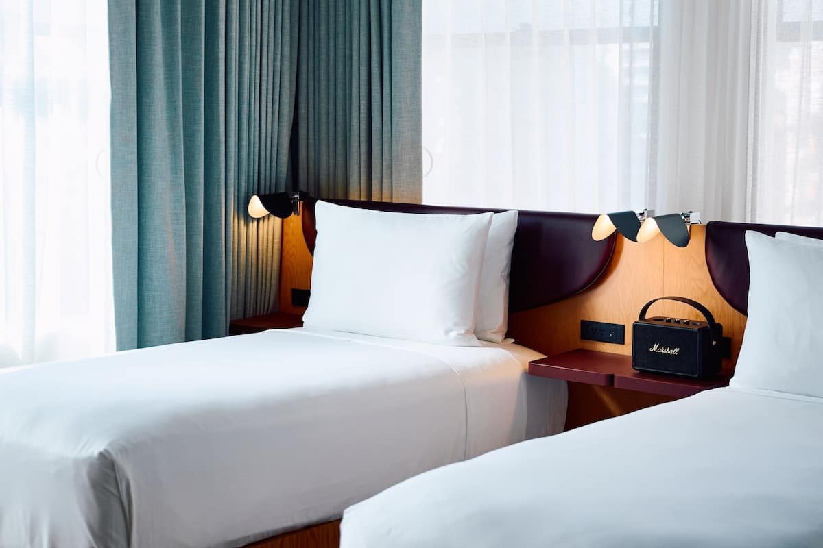 Hotellihuone, jossa on kaksi sänkyä, yöpöytä lampun kanssa, radio ja läpikuultavat verhot.