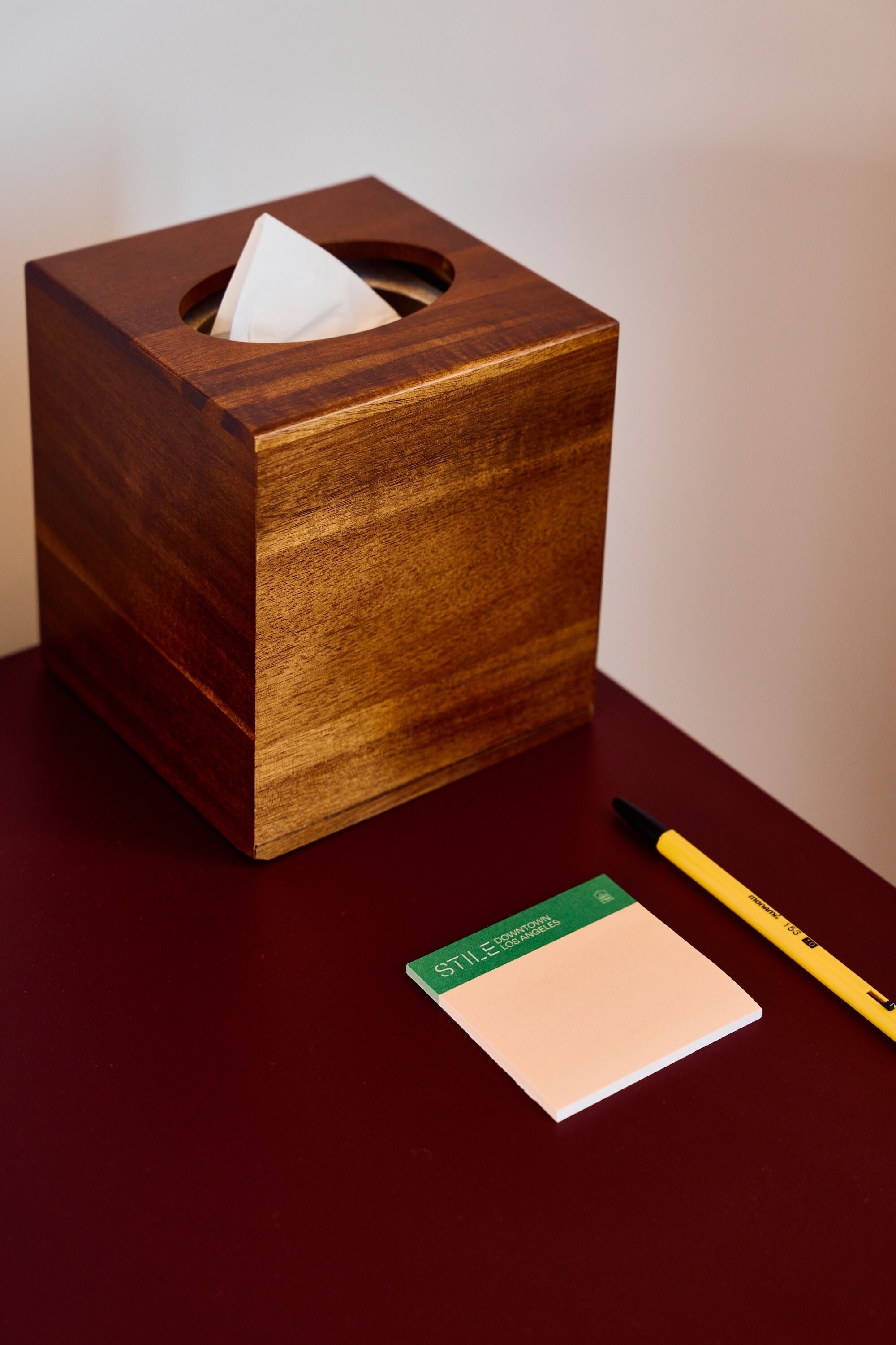 Een houten tissuedoos met daarin een tissue, een notitieboekje en een gele pen op een donker oppervlak.