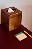 Una caja de pañuelos de madera con un pañuelo dentro, un bloc de notas y un bolígrafo amarillo sobre una superficie oscura.