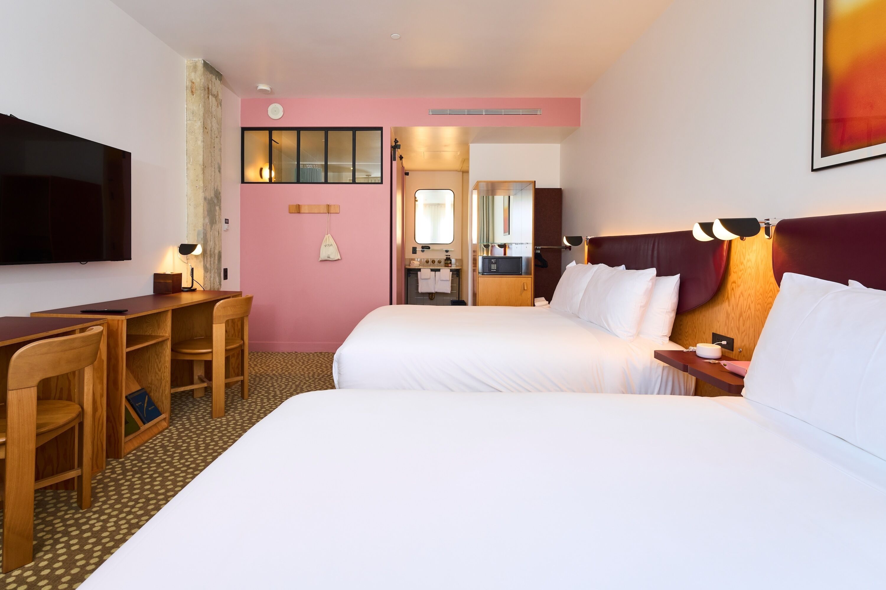 A hotel room with two beds, a desk, chairs, a TV, a mirror, and a bathroom visible through an open door.