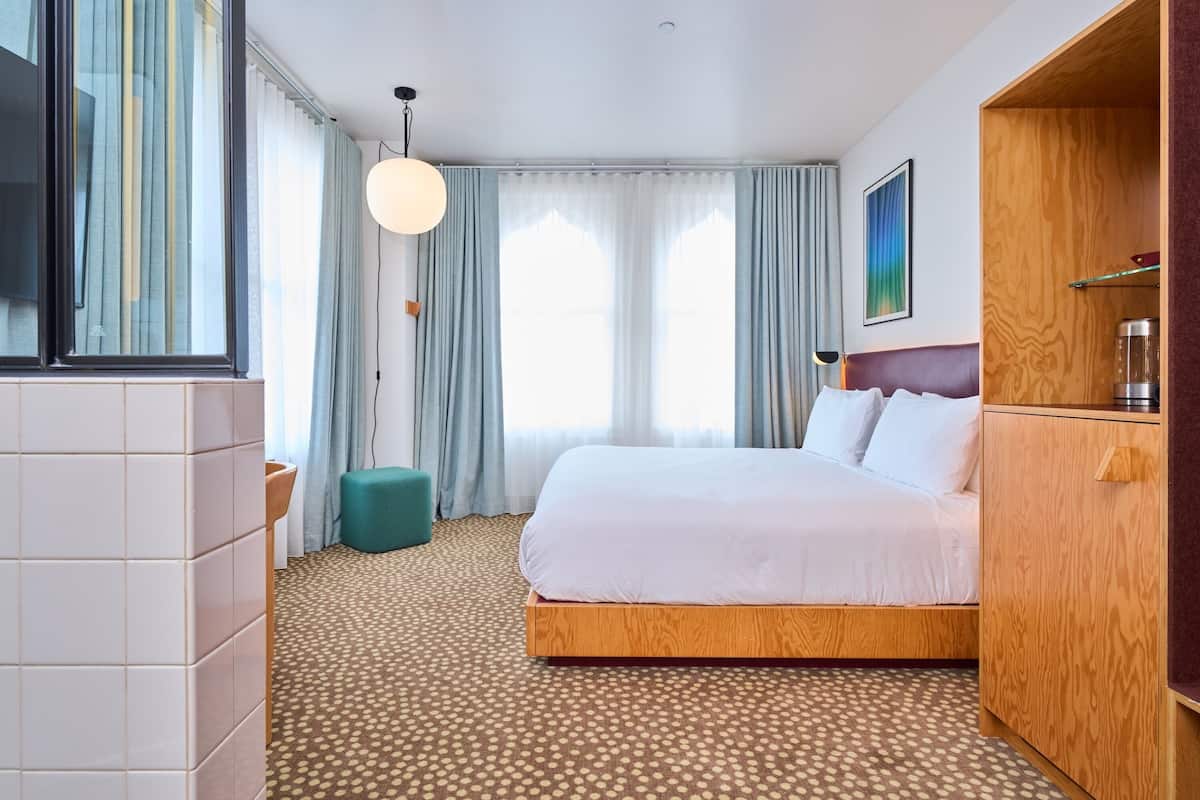 Hotellihuone, jossa on sänky, puinen pääty, suuri ikkuna verhoilla, pyöreä valaisin ja laatoitettu lattia.